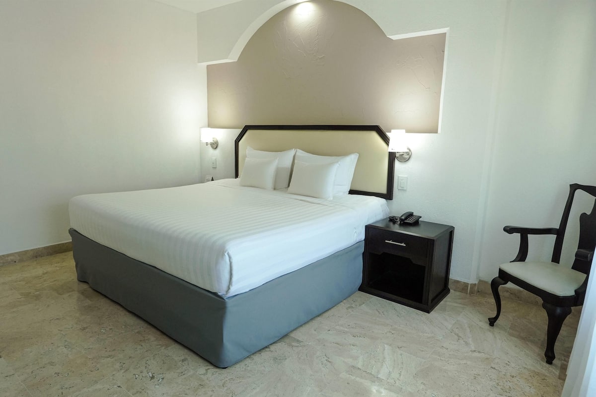A neatly arranged hotel room with a large bed, bedside table, chair, and a wall-mounted light fixture.