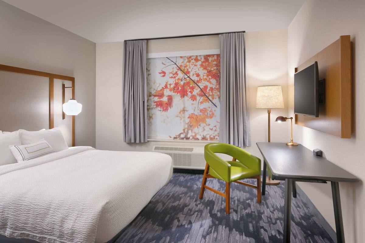 Una habitación de hotel con cama, escritorio, silla, televisor y ventana con cortinas.