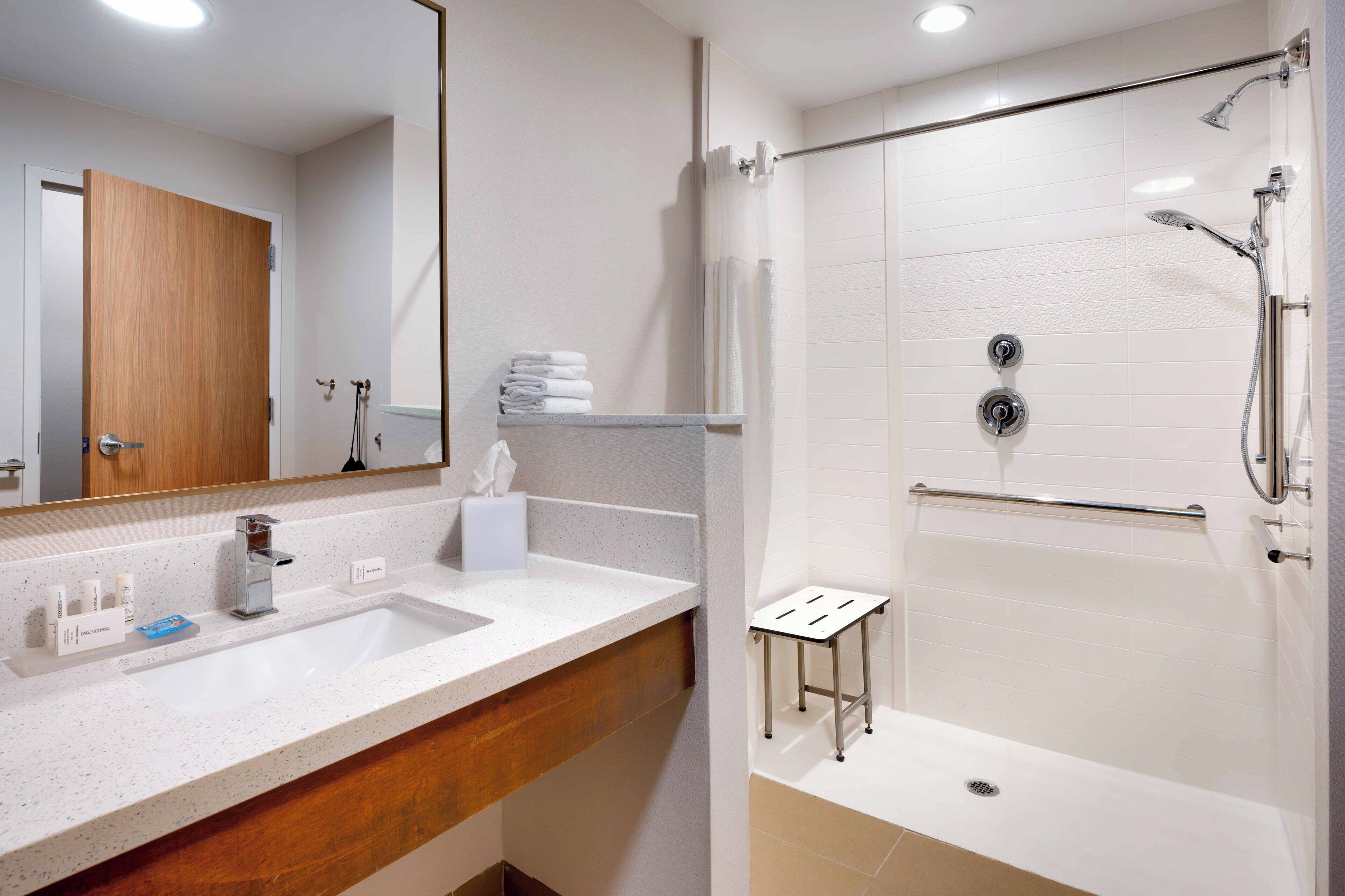 A modern bathroom with a large mirror, a sink with toiletries, a shower with a handheld showerhead, and a towel rack.
