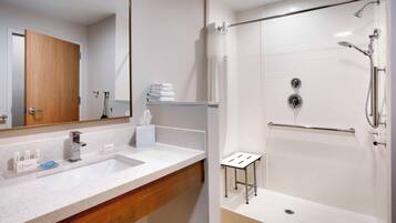 A modern bathroom with a large mirror, a sink with toiletries, a shower with a handheld showerhead, and a towel rack.