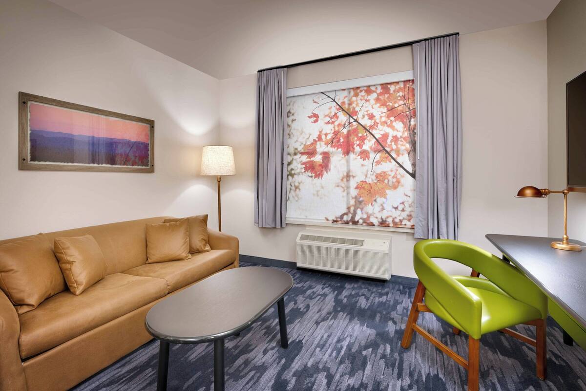 Una habitación con un sofá marrón, una pequeña mesa redonda, una silla verde, una lámpara y un ventanal grande con cortinas.