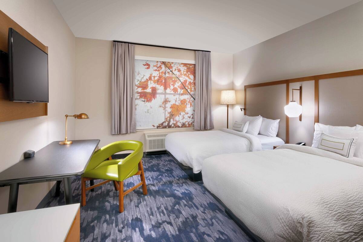 Habitación de hotel con cama, escritorio, silla, televisor y una ventana grande con cortinas.