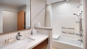 A modern bathroom with a large mirror, a sink with toiletries, a shower with a handheld showerhead, and a bathtub.