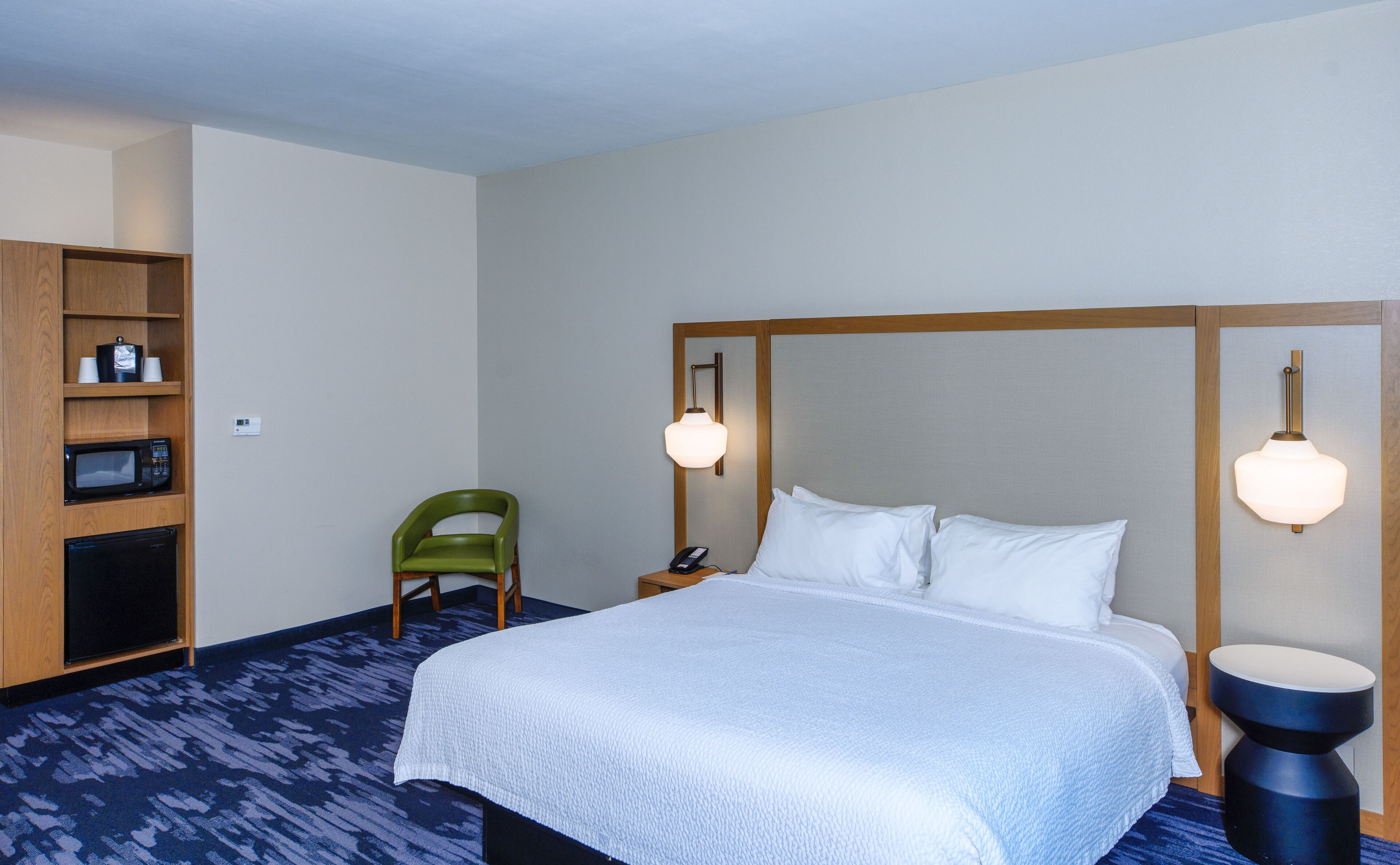 Ein Hotelzimmer mit einem großen Bett, einem Holzkopfende, einem grünen Sessel, einer Mikrowelle, einem Kühlschrank und einem Regal mit Tassen und einem Wasserkocher.