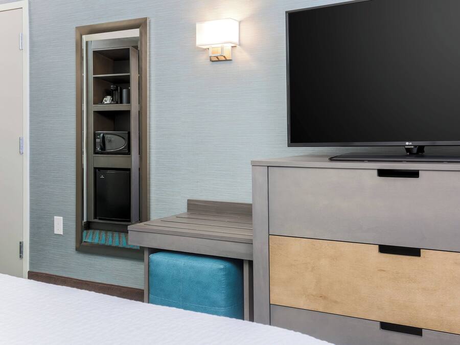 Una habitación de hotel moderna con televisión de pantalla plana, estanterías empotradas con microondas y una banca.