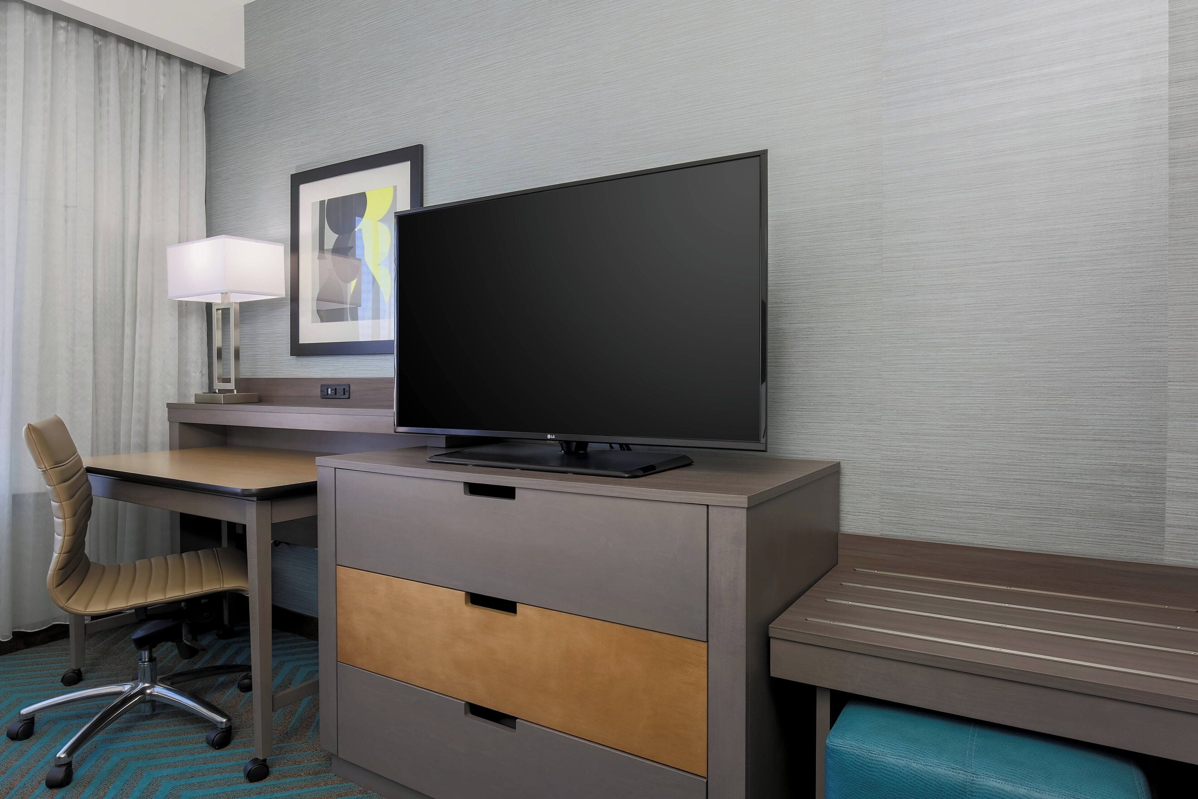A hotel room with a desk, chair, TV, lamp, and artwork on the wall.