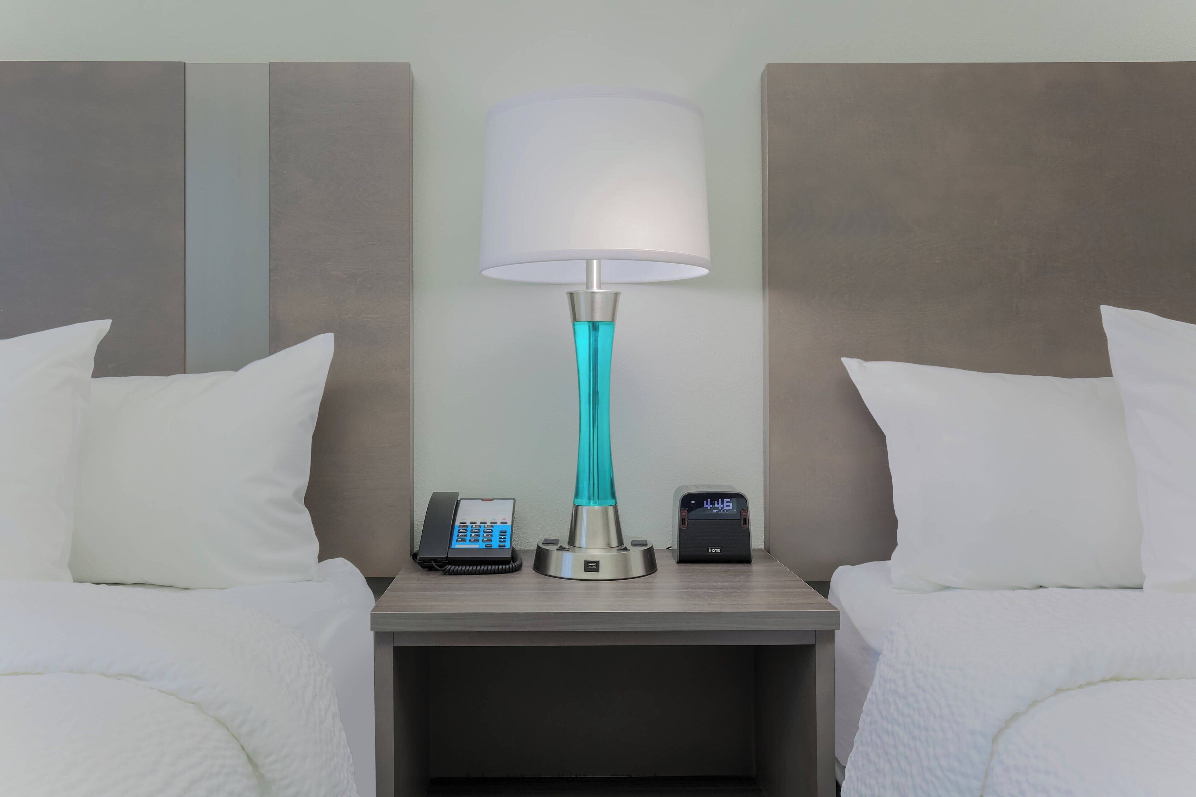 A hotel room with a bed, a nightstand with a lamp, a phone, and a digital clock.