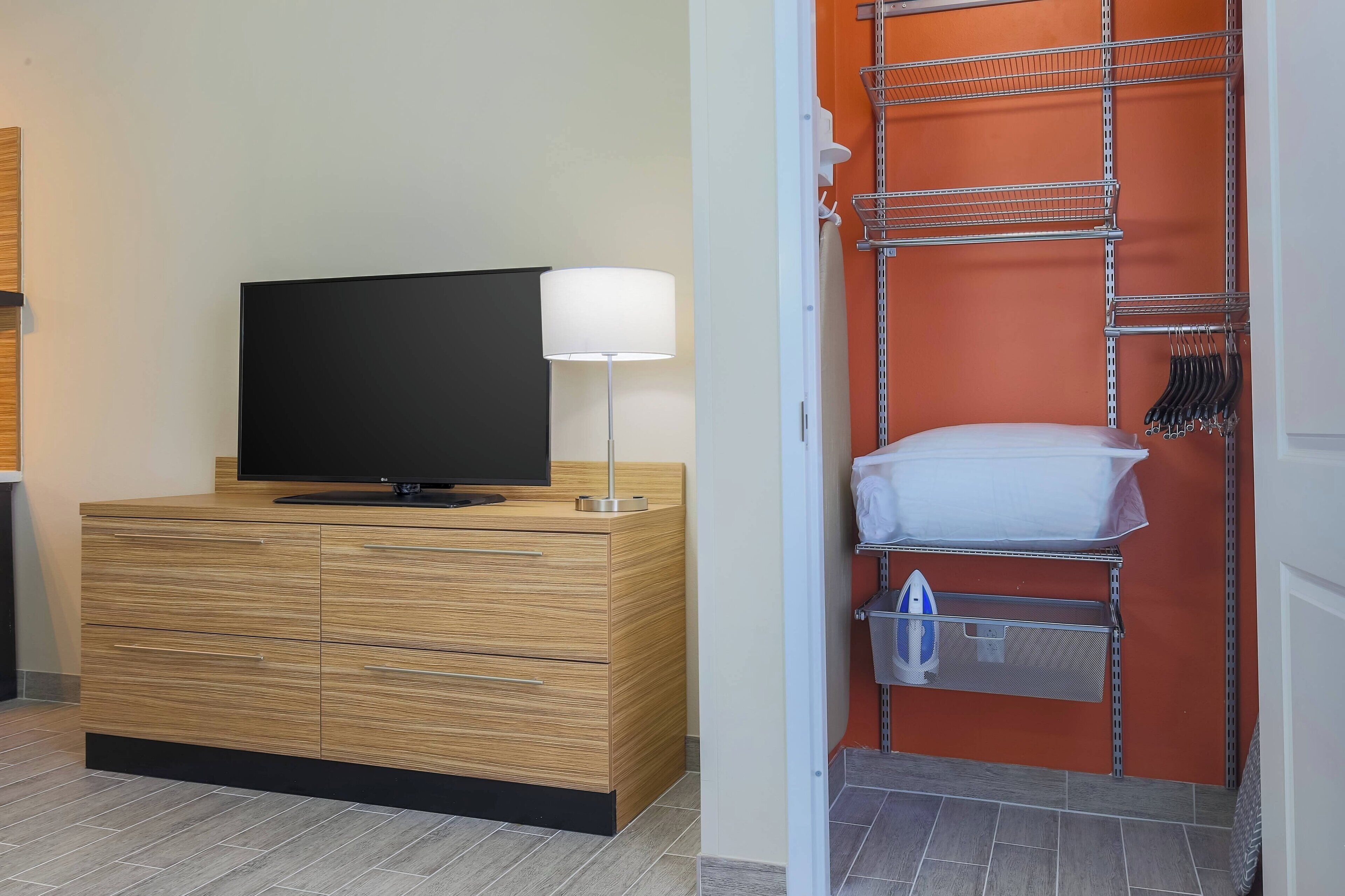 A modern hotel room with a wooden dresser, a flat-screen TV, a white lamp, and a metal shelving unit with hanging clothes.