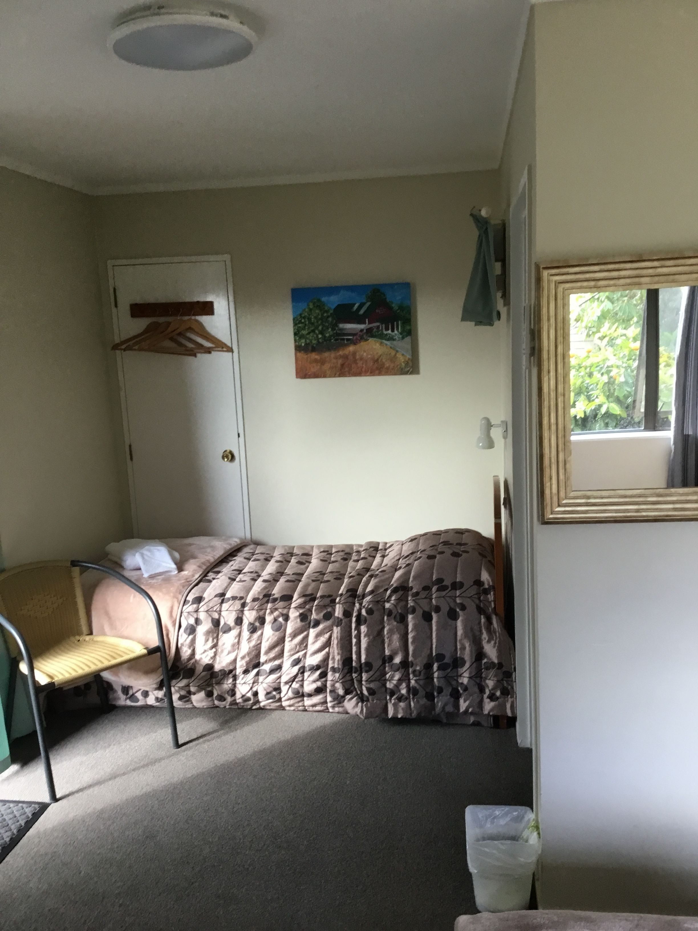A small, single-bed room with a bed, a chair, a door, a painting on the wall, and a window.