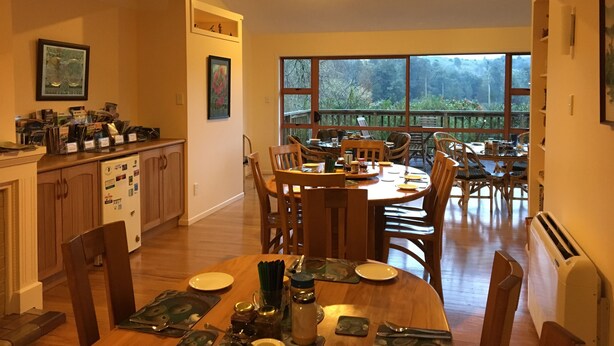 A dining area with a wooden table and chairs, a kitchen with cabinets and a refrigerator, and a large window offering a view of the outdoors. - Waitomo Caves Guest Lodge (Waitomo Caves)