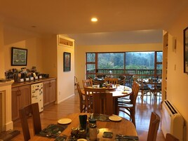 Uma área de refeições com mesa e cadeiras de madeira, uma cozinha com armários e geladeira, e uma grande janela com vista para o exterior.