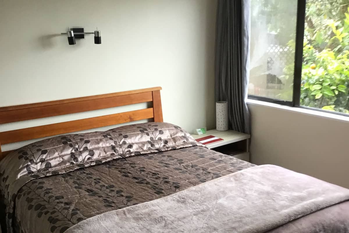 Ein ordentlich gemachtes Bett mit einem blumigen Bettüberwurf und einer grauen Decke, ein hölzernes Kopfteil, ein kleiner Nachttisch mit einer Lampe und ein Fenster mit Blick ins Grüne.