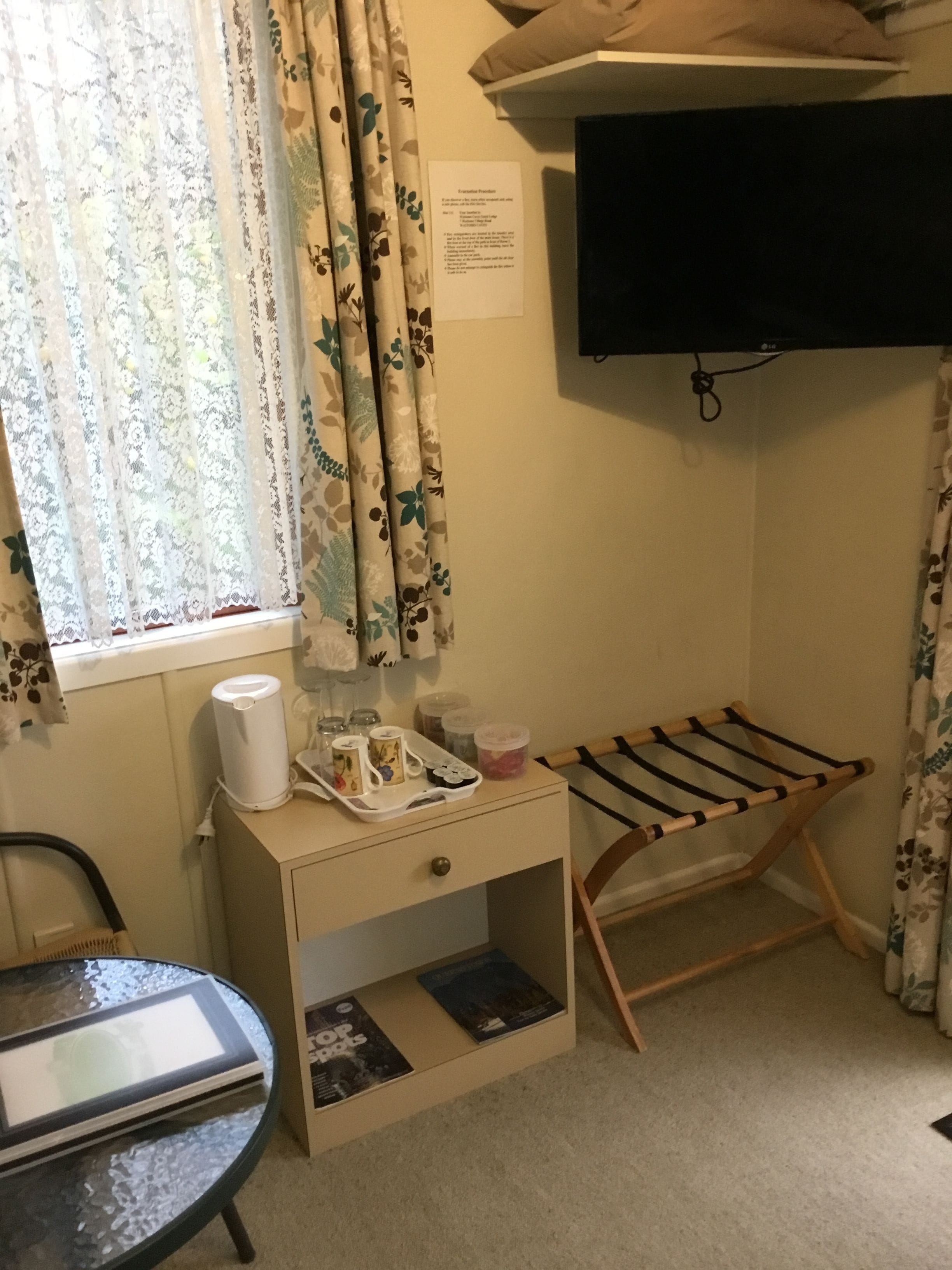 A room with a window, a flat-screen TV mounted on the wall, a small wooden side table with a kettle and a tray, and a glass-top table with magazines.