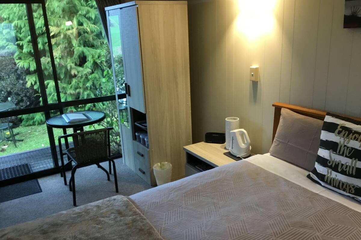 Ein Hotelzimmer mit einem Bett, einem kleinen Tisch, einem Sessel, einem Nachttisch, einer Lampe und Blick auf Grünfläche.