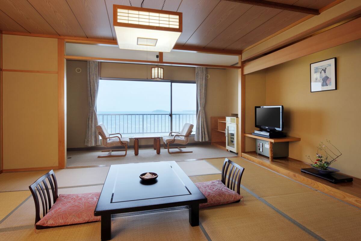 A traditional Japanese room with tatami flooring, a low table with cushions, a television, and a view of the sea.