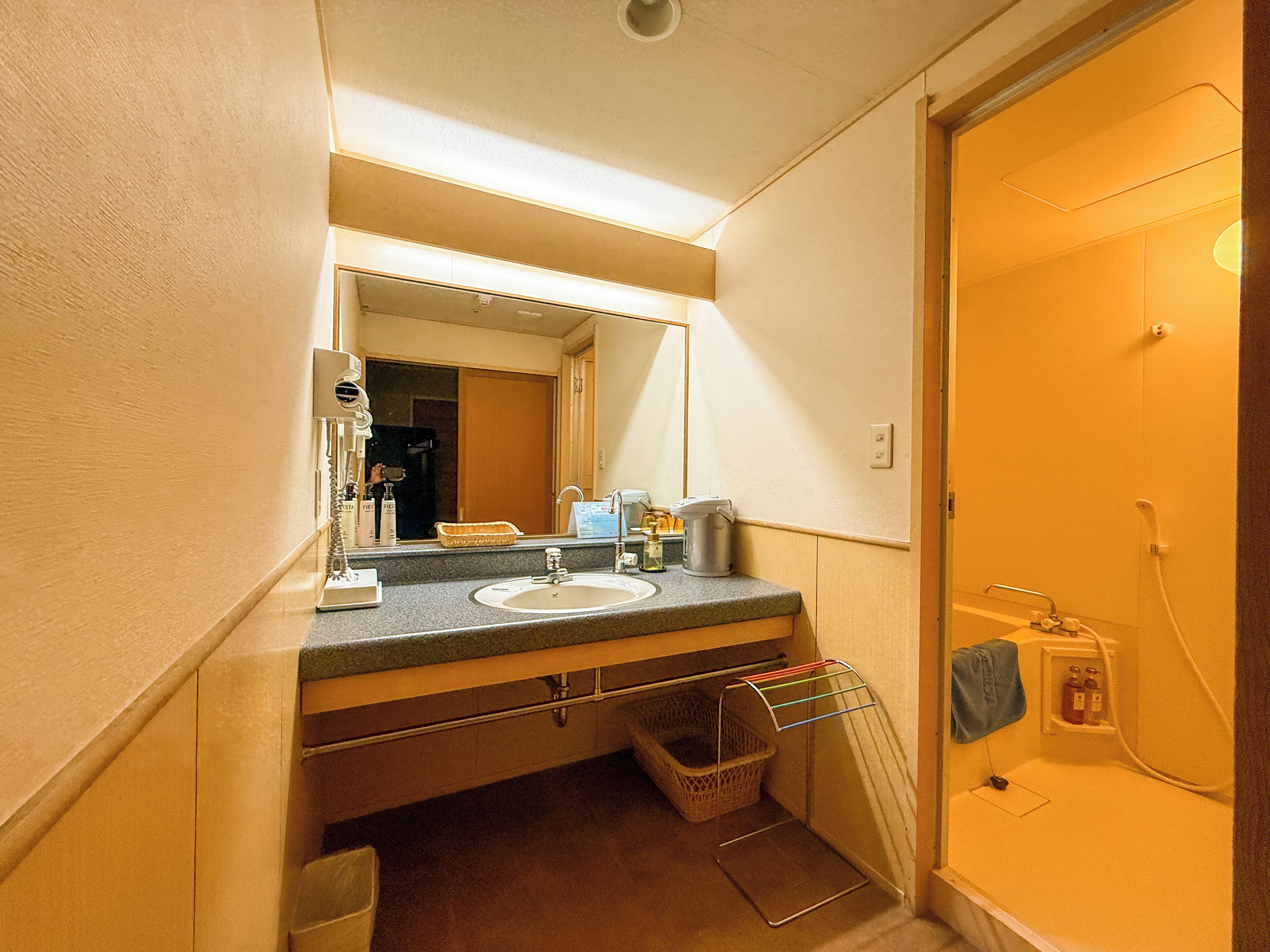 Un baño con lavamanos, espejo y ducha. Hay un perchero y una cesta debajo del lavamanos.