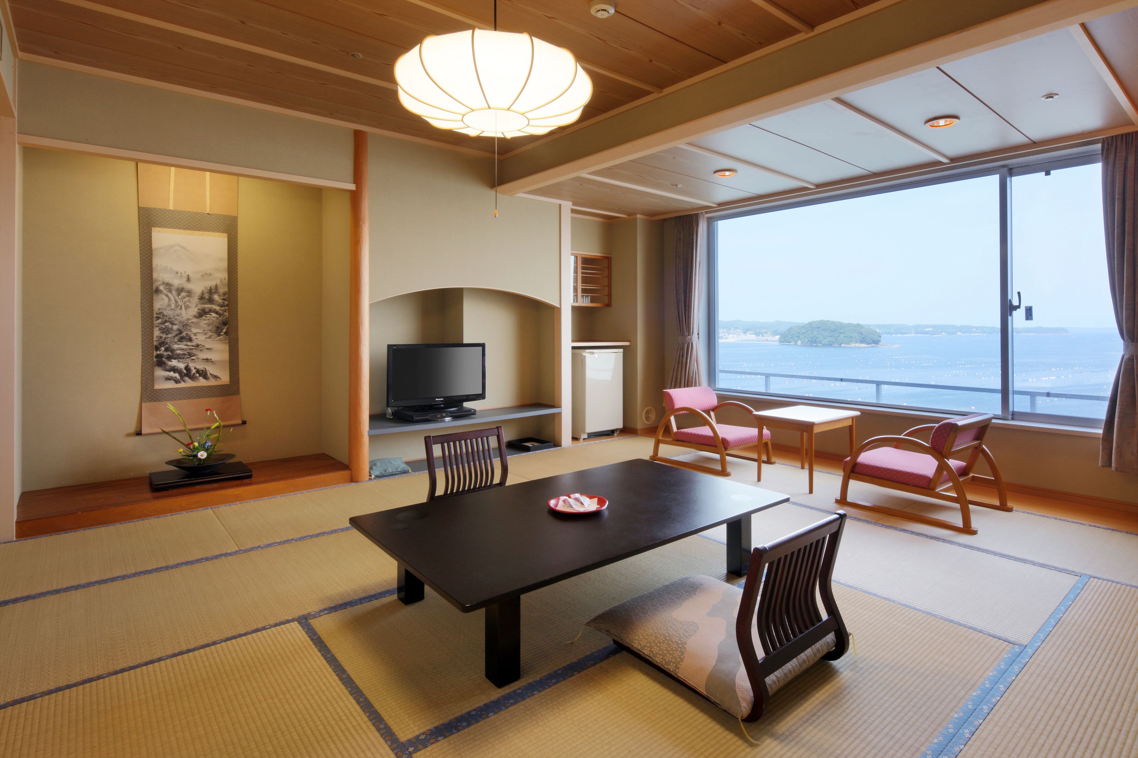 A traditional Japanese room with tatami flooring, a low table, a television, and a view of the sea.