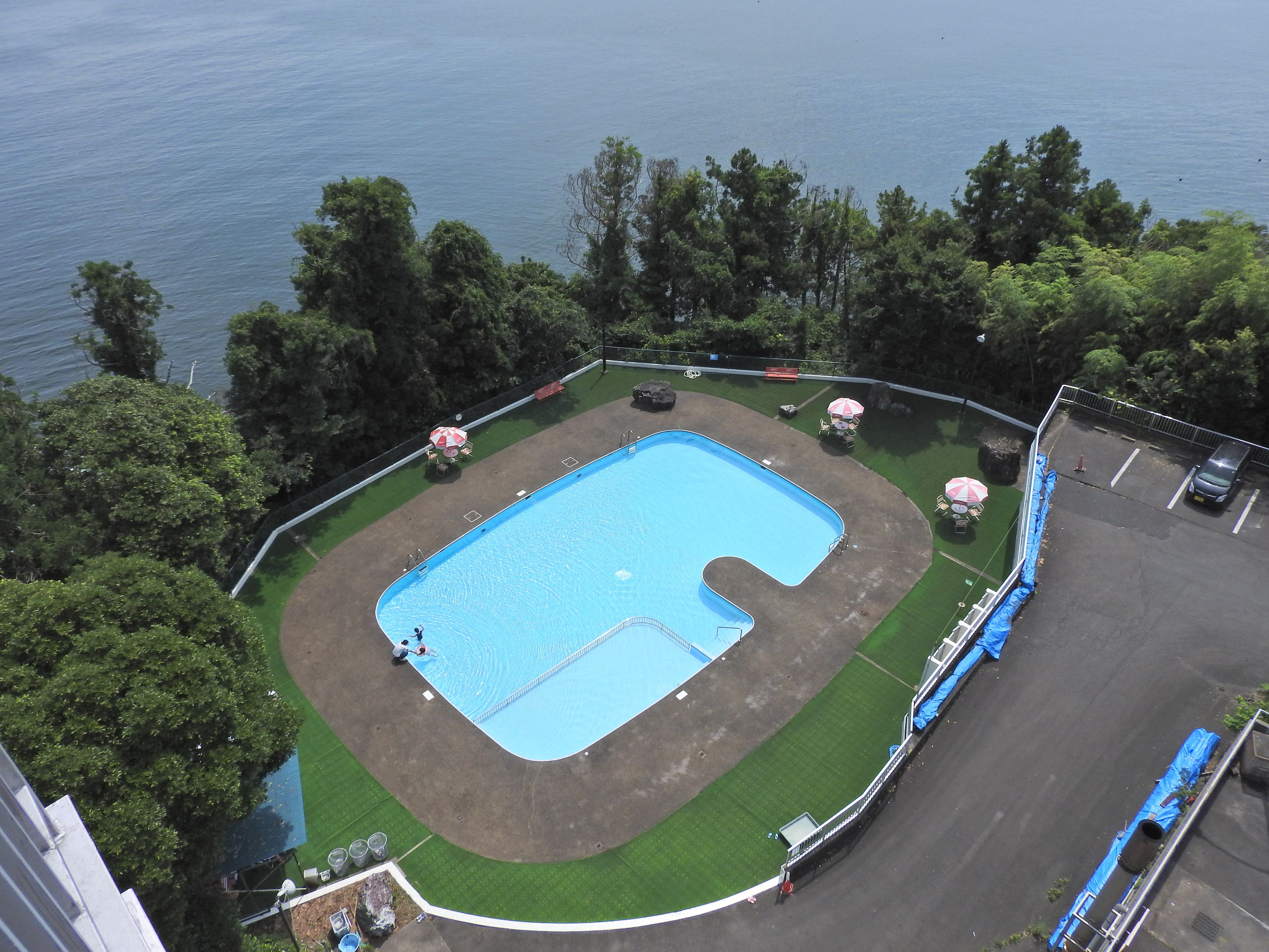 An aerial view of a swimming pool surrounded by a fenced area with trees and a parking lot.