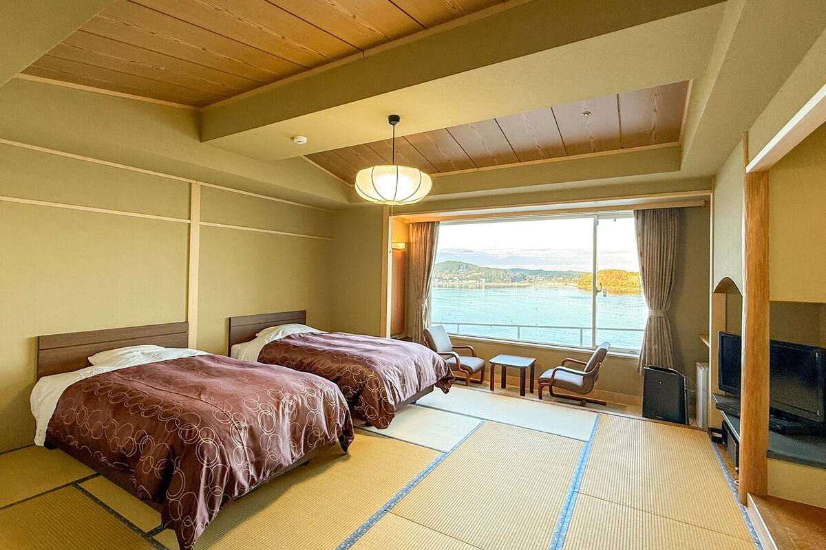 A hotel room with two beds, a view of a body of water, a television, and a traditional Japanese tatami mat floor.