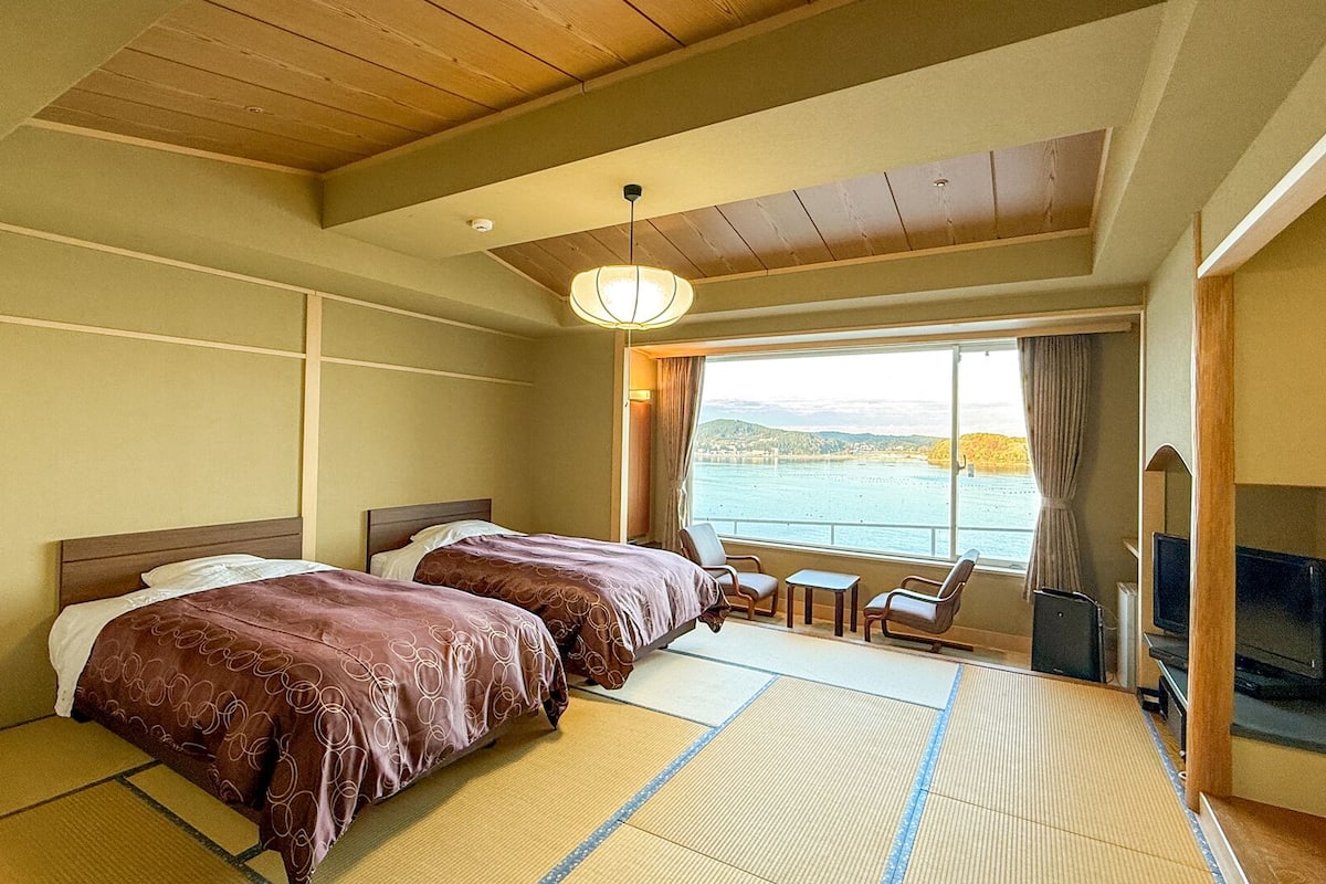 A hotel room with two beds, a view of a body of water, a television, and a traditional Japanese tatami mat floor.