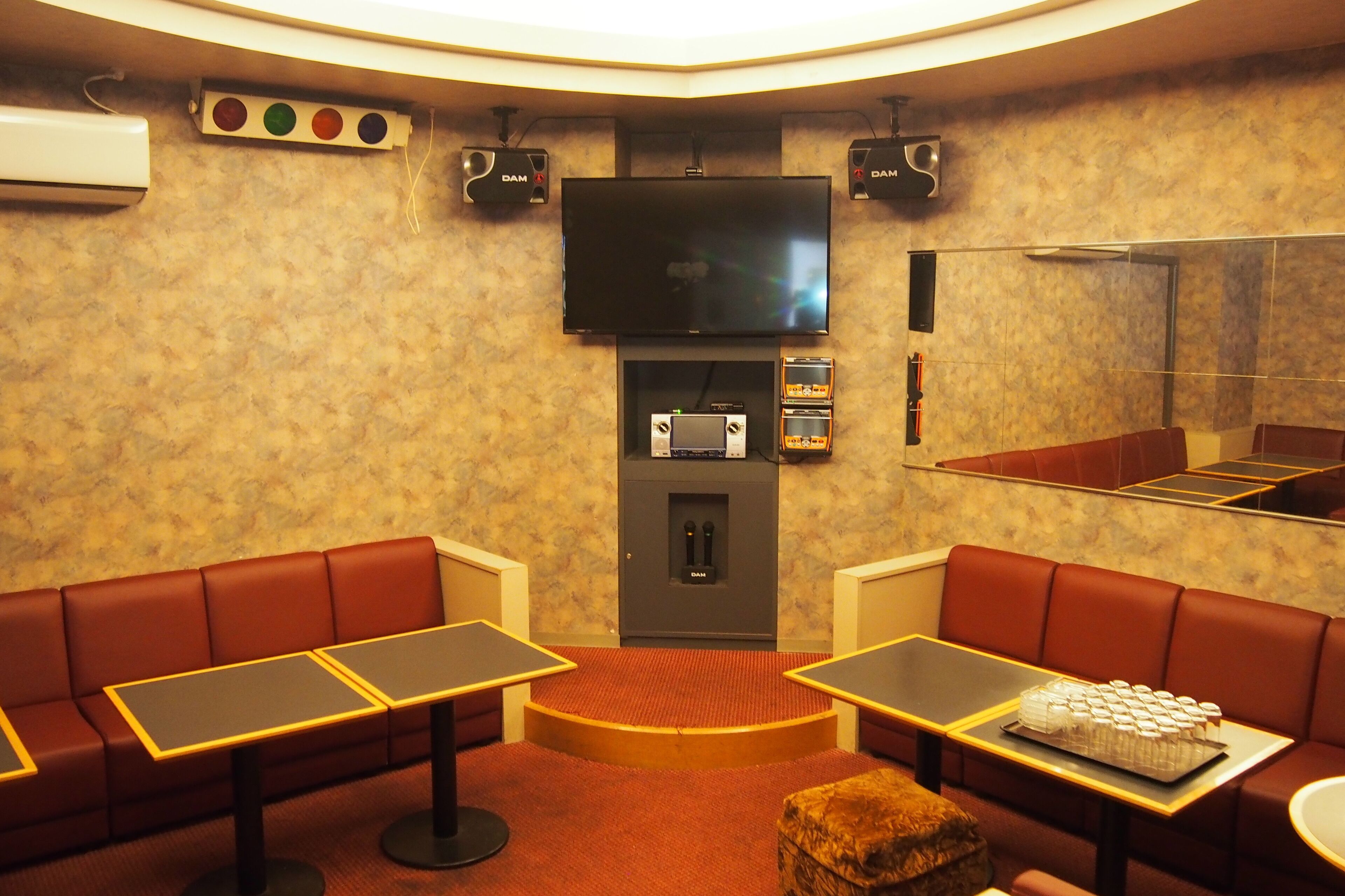 A room with a flat-screen TV mounted on the wall, a sound system, a small refrigerator, and a seating area with tables and benches.