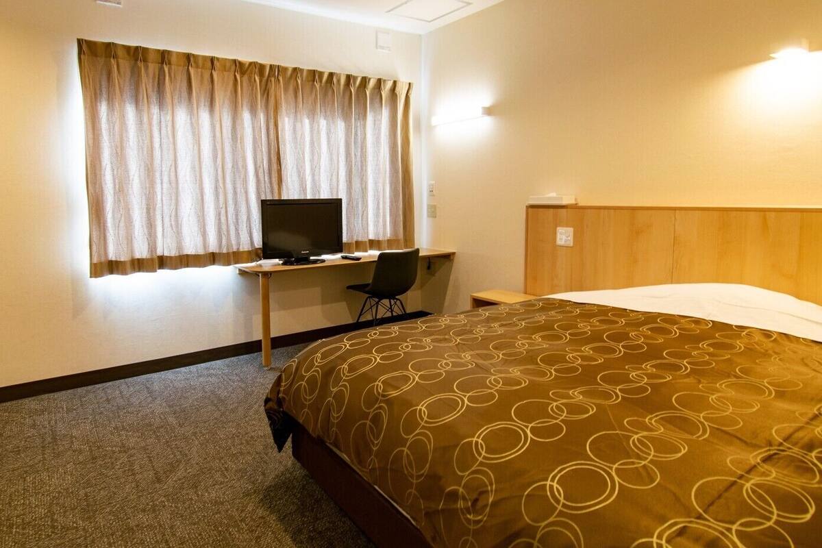 A hotel room with a bed, a desk with a computer, a chair, and a window with curtains.