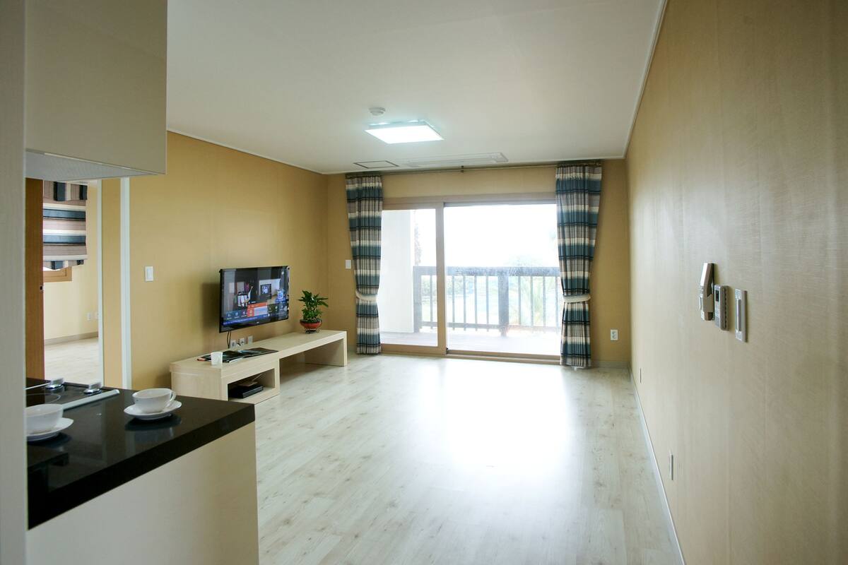 A modern living room with a flat-screen TV, a small table, and a bench. There is a sliding glass door leading to a balcony.