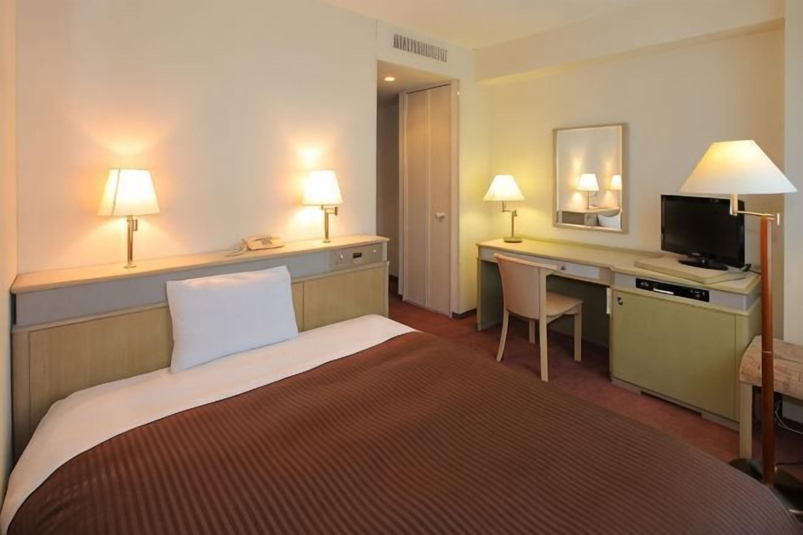 Una habitación de hotel con una cama, lámparas de noche, un escritorio con silla, un televisor y un espejo.