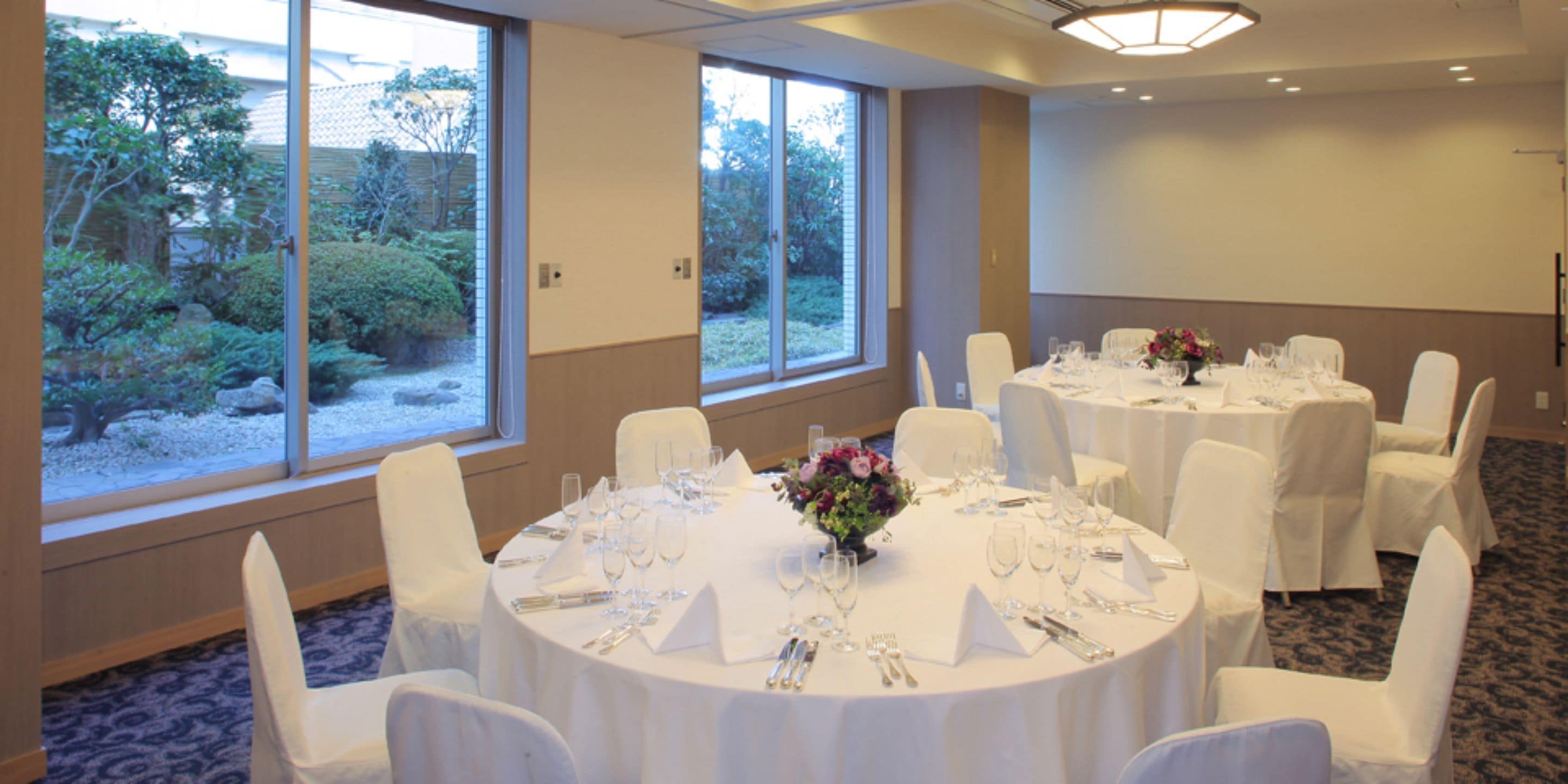 Un salón de conferencias con mesas redondas dispuestas para un evento formal, amplias ventanas con vistas a zonas verdes y suelo alfombrado.