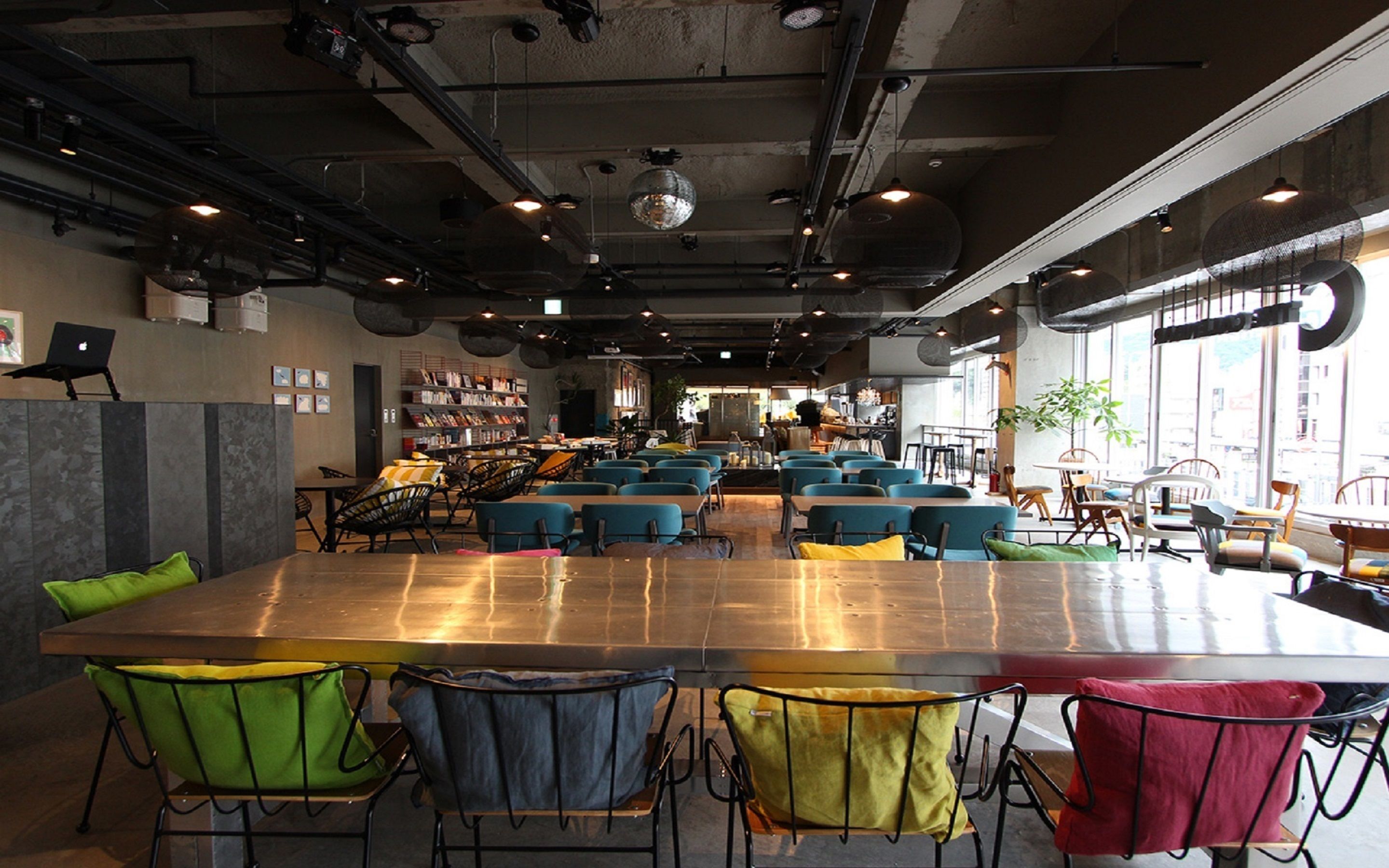 A modern cafe with a long communal table, multiple seating areas, and a visible kitchen in the background.