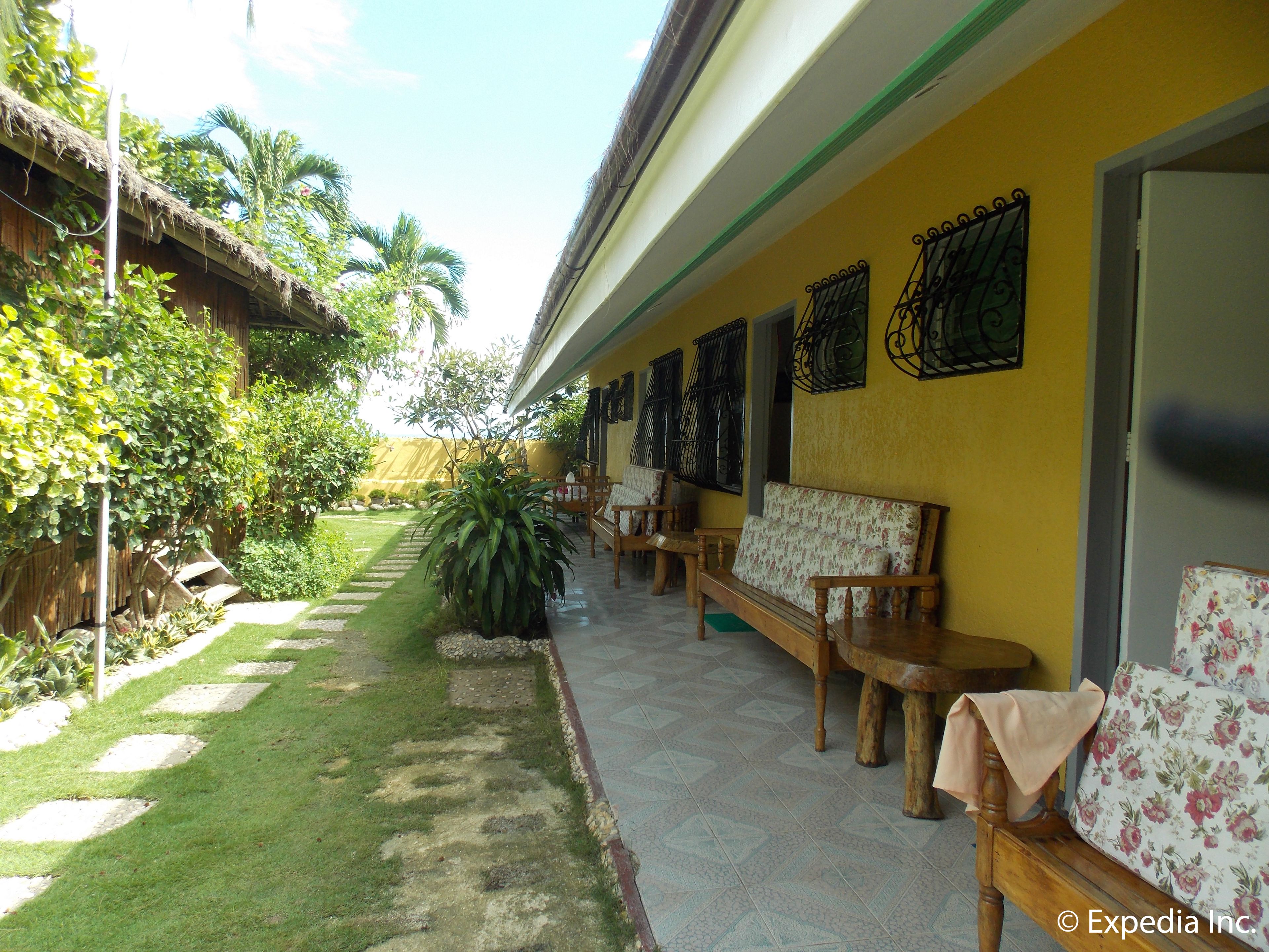 A covered outdoor seating area with wooden benches and tables, surrounded by a garden with a stone pathway.