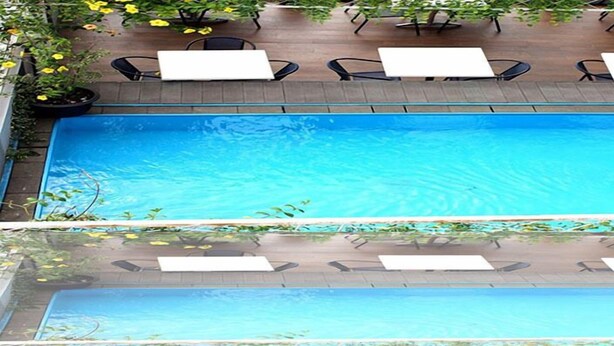 Ein rechteckiger Pool im Freien mit klarem, blauem Wasser, umgeben von einer Holzterrasse und Sitzgelegenheiten mit weißen Tischen und schwarzen Stühlen.