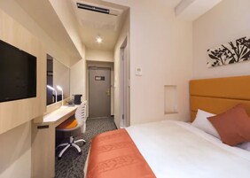 Quarto de hotel com cama, escrivaninha com cadeira, televisão e porta para outro espaço.