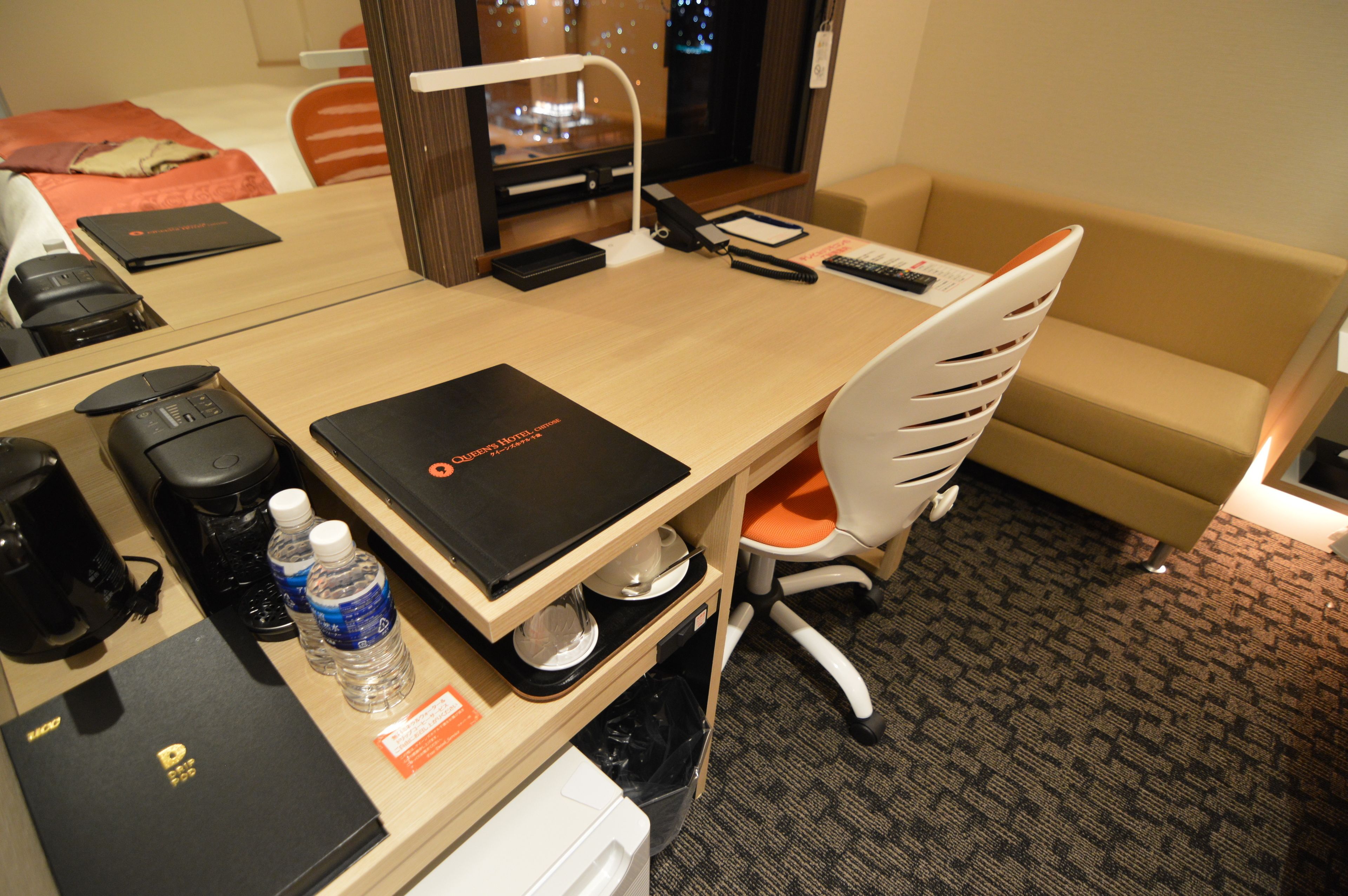 Ein Hotelzimmer mit einem Holztisch, einer schwarzen Ordner, einer Wasserflasche, einer Kaffeemaschine, einer weißen Mülltonne und einem beigen Sofa.