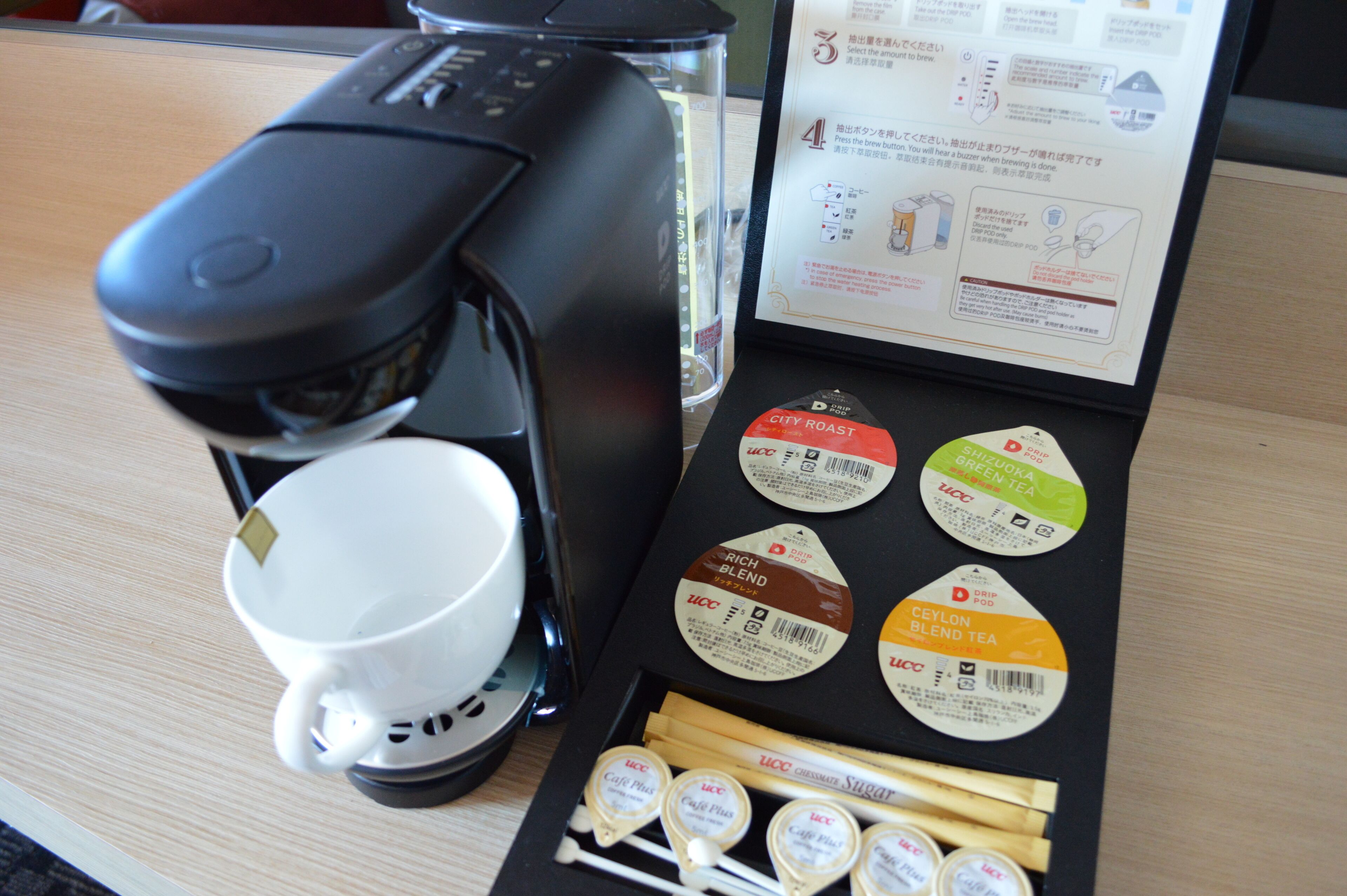 Une machine à café, une tasse et une boîte de différents mélanges de thé sont posés sur une table.