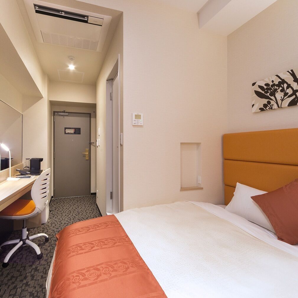 A hotel room with a bed, a desk with a chair, a mirror, a door, and a wall-mounted light fixture.