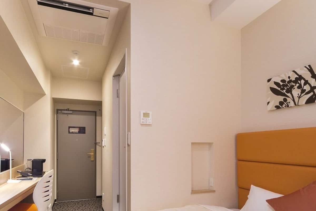 Habitación de hotel con cama, un escritorio y silla, un espejo, una puerta y una lámpara fijada a la pared.