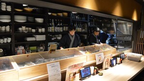 A sushi bar with a wooden counter, clear plastic containers, and a menu stand. - Queen's Hotel Chitose (Chitose)
