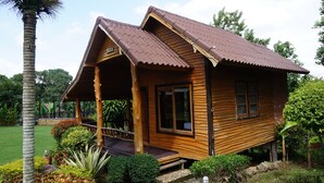 Una cabaña de madera con techo inclinado, un porche y un pequeño jardín con diversas plantas y una palmera.