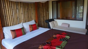 Una cama con colcha marrón y almohadas rojas y verdes, pared de madera y ventana con persianas.