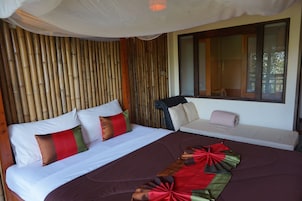 A bed with a brown bedspread and red and green pillows, a wooden wall, and a window with blinds.