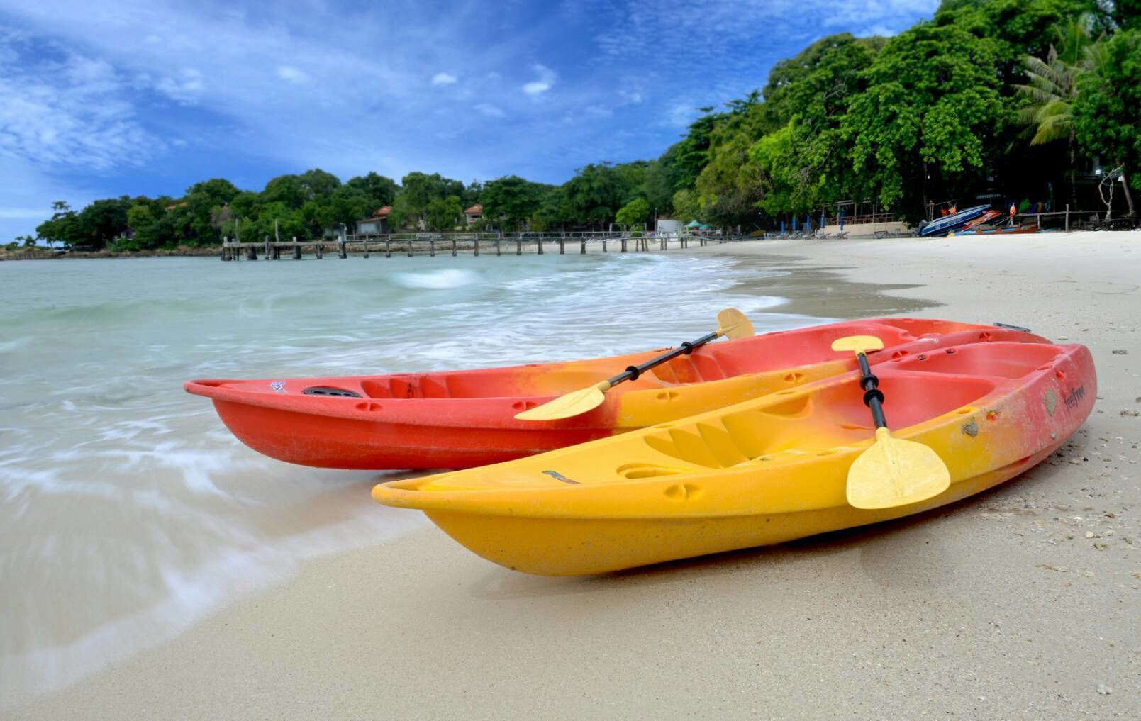 Two kayaks on a sandy beach with a wooden pier and lush greenery in the background.