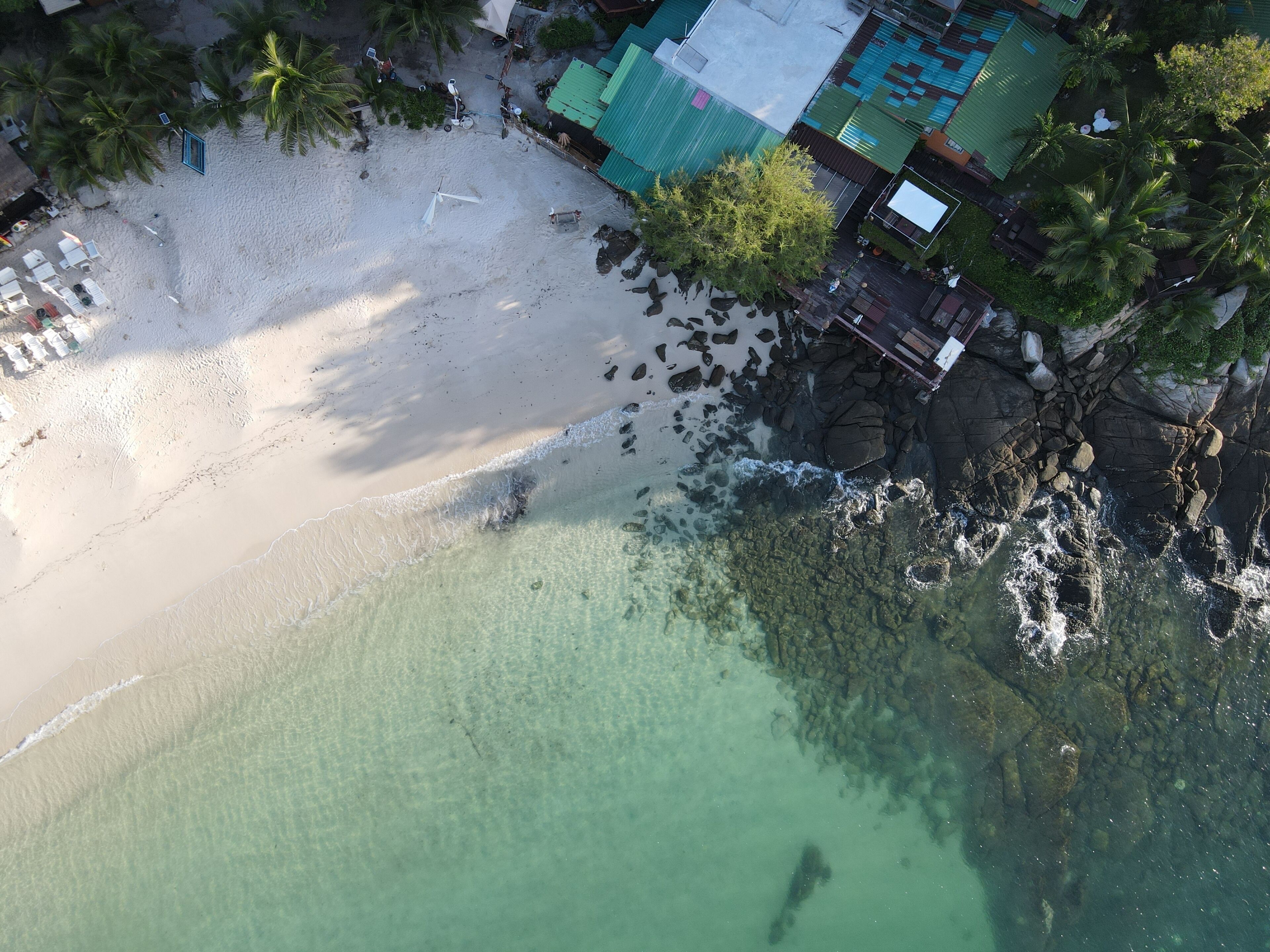 An aerial view of a beach with clear turquoise water, a sandy shore, and a building with a green roof.