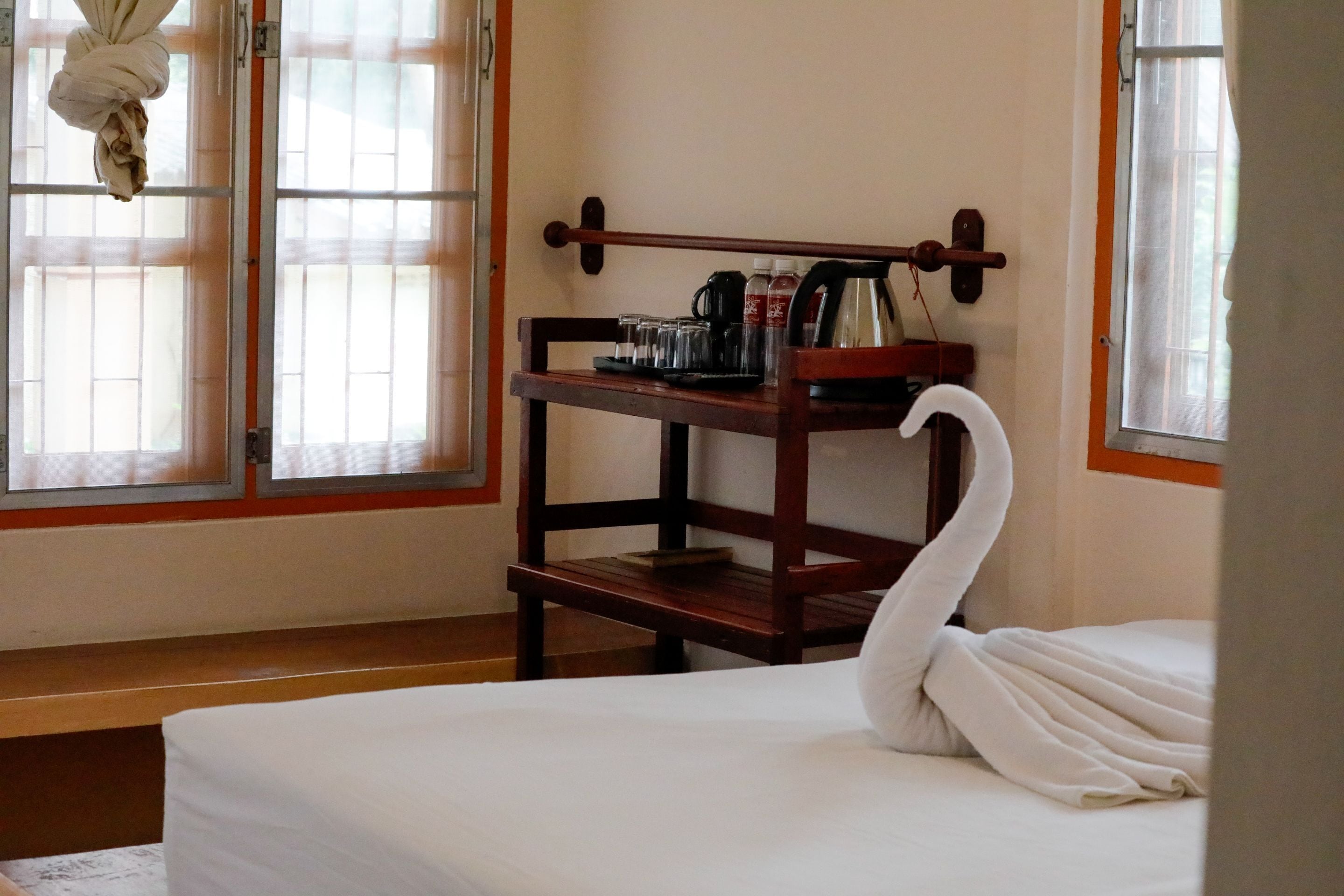 Una camera d'albergo con un letto, un comodino in legno con bollitore, tazze e una bottiglia, e una finestra con tende.