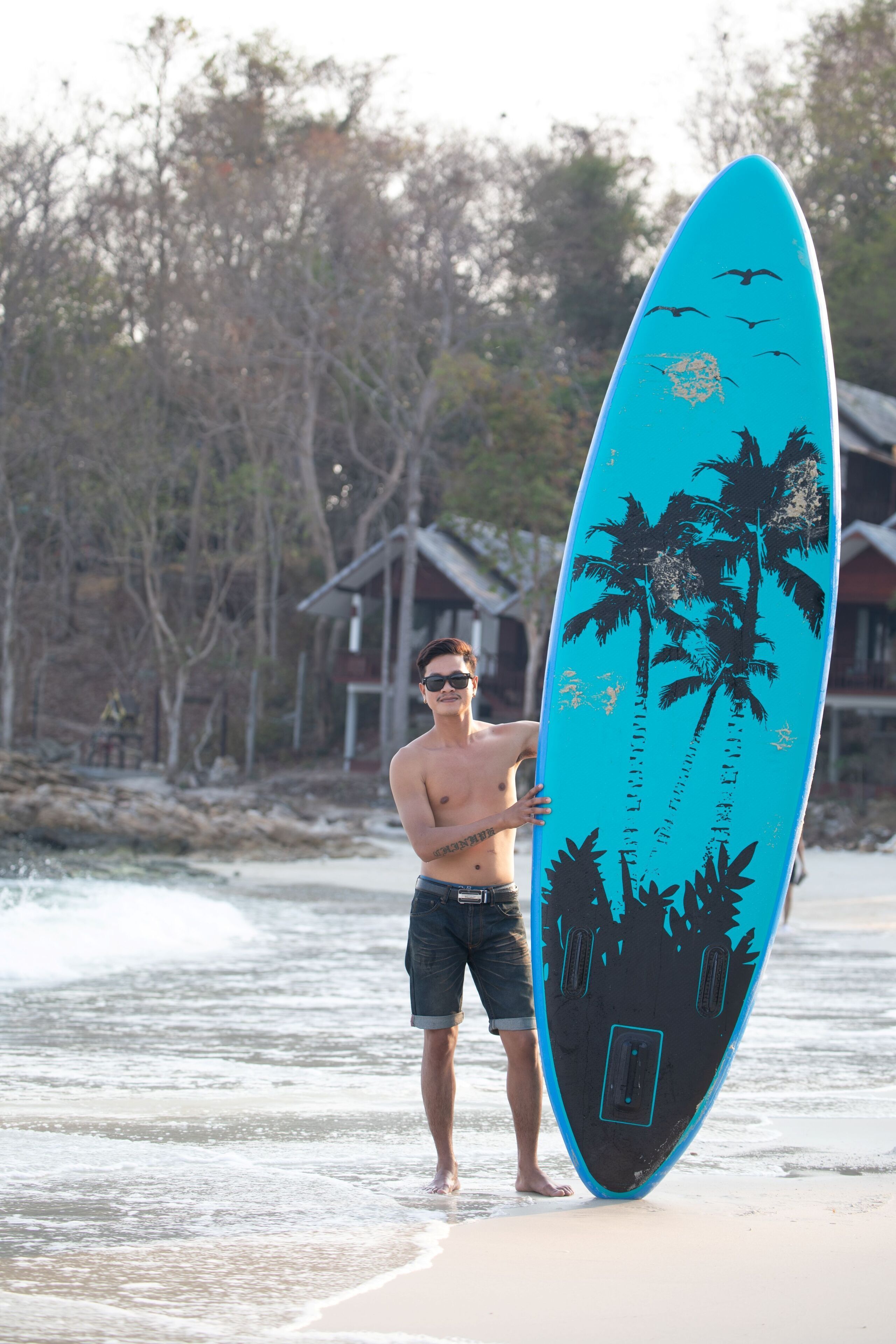 A person holding a surfboard on a beach with palm trees and houses in the background.