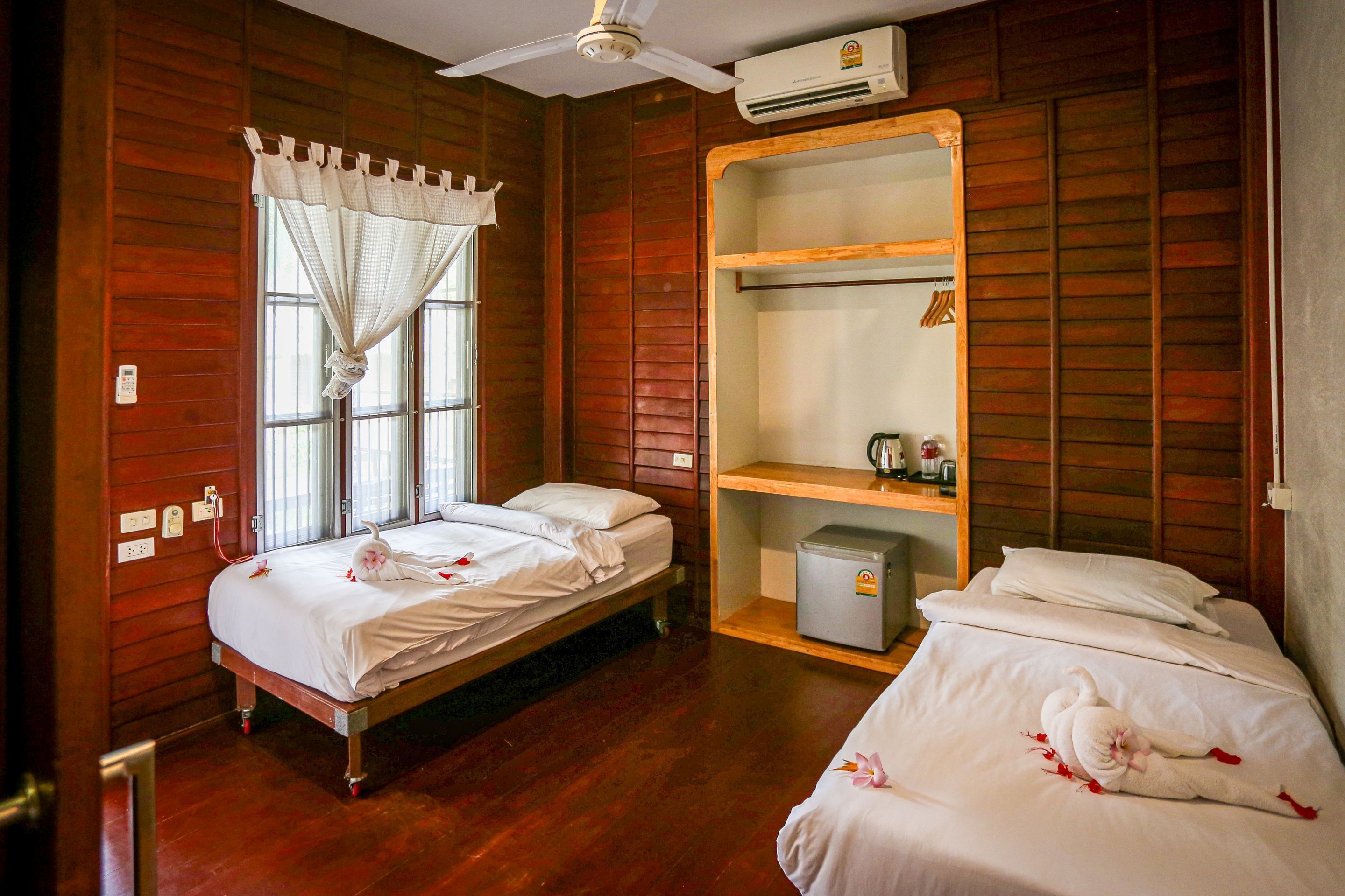 A room with two beds, wooden walls, a ceiling fan, an air conditioner, a window with curtains, and a shelf with a kettle.
