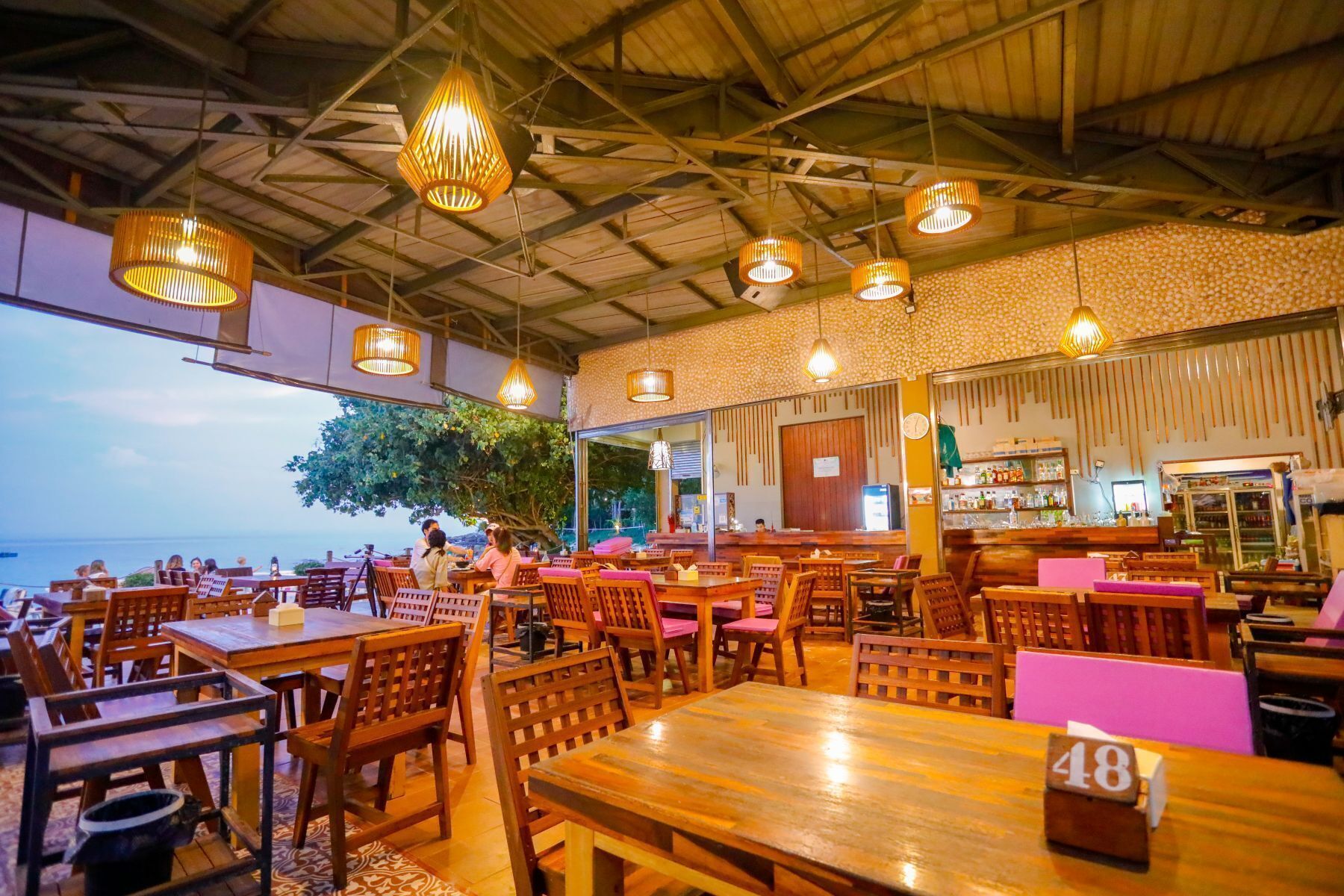 Un restaurante con mesas y sillas de madera, luces colgantes y vista al mar.