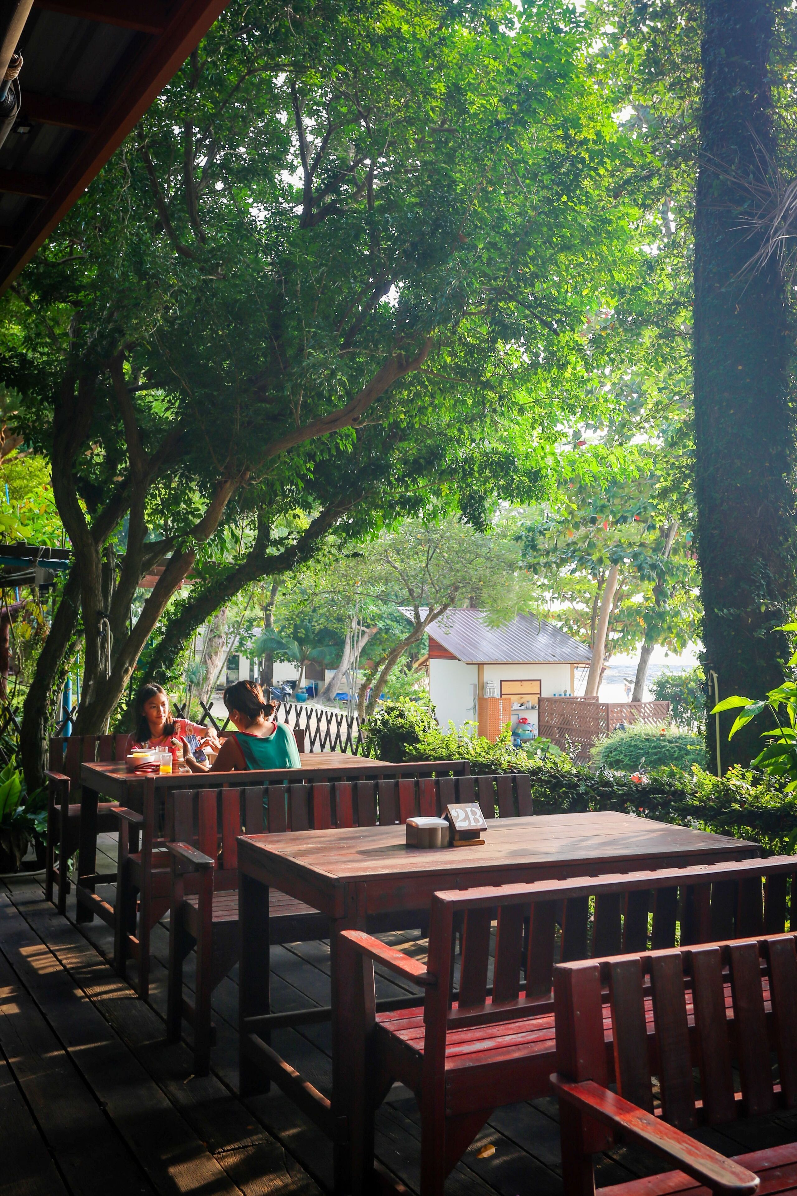 An outdoor seating area with wooden tables and chairs, surrounded by lush greenery and trees.