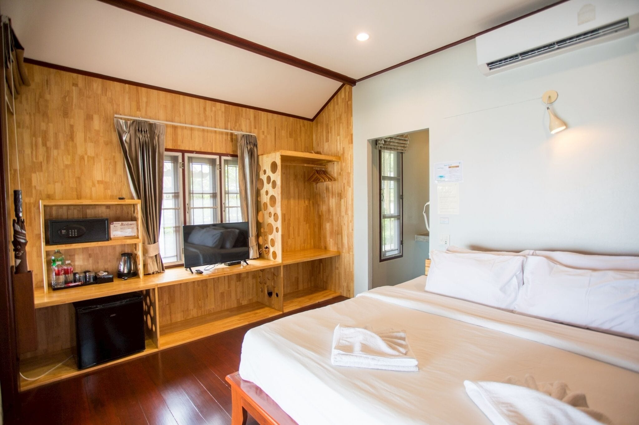 A hotel room with a wooden wall, a bed, a TV, a mini fridge, and a desk with a computer and a lamp.