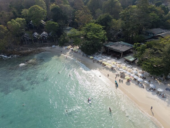 Vista aérea de una playa con agua turquesa cristalina, una orilla arenosa y una fila de sombrillas y sillas de playa.